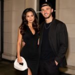 Kennedy Alexa wearing a black dress posing elegantly next to her partner, footballer James Maddison, in a matching black outfit and cap.