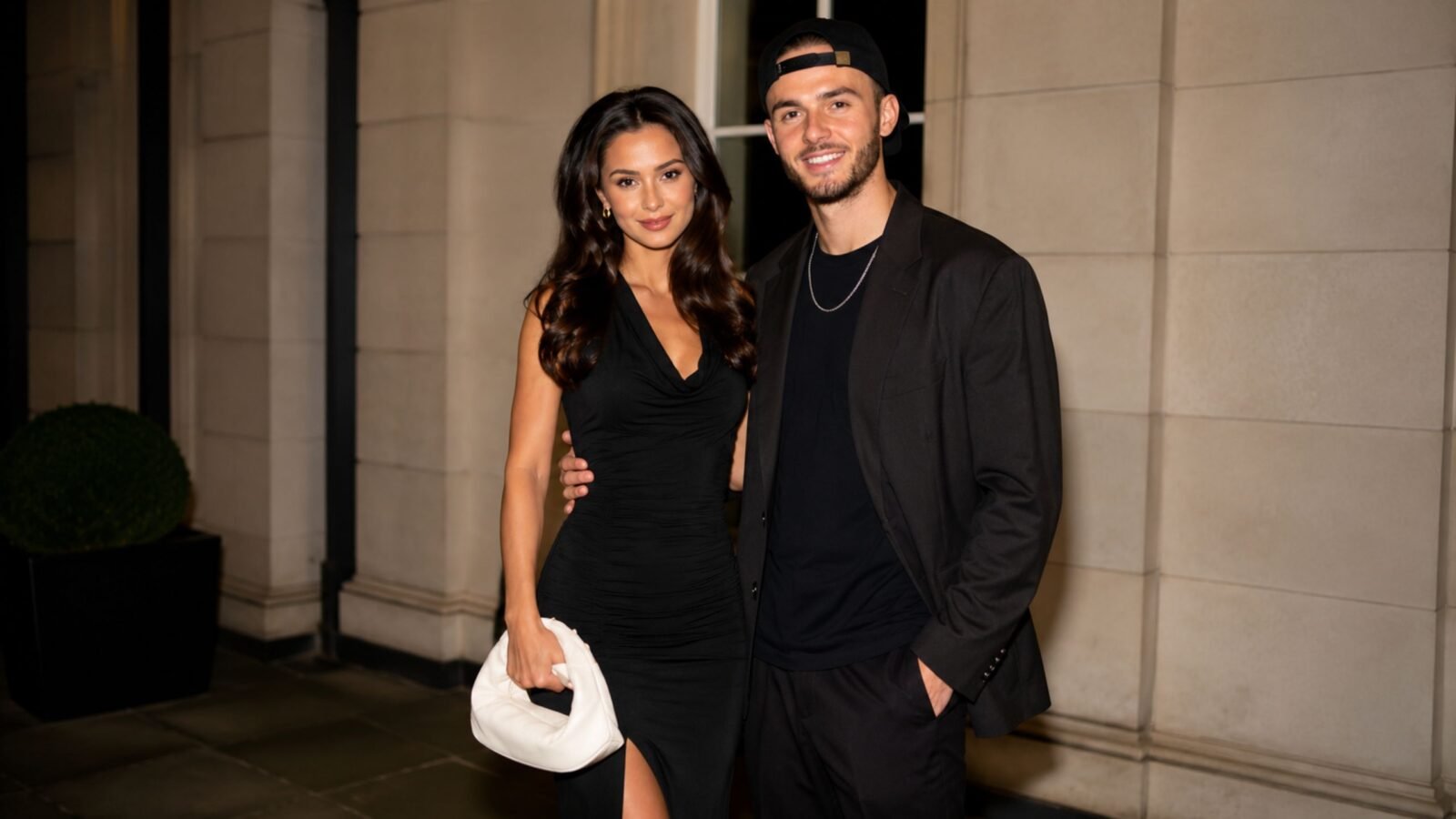 Kennedy Alexa wearing a black dress posing elegantly next to her partner, footballer James Maddison, in a matching black outfit and cap.