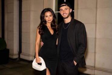 Kennedy Alexa wearing a black dress posing elegantly next to her partner, footballer James Maddison, in a matching black outfit and cap.
